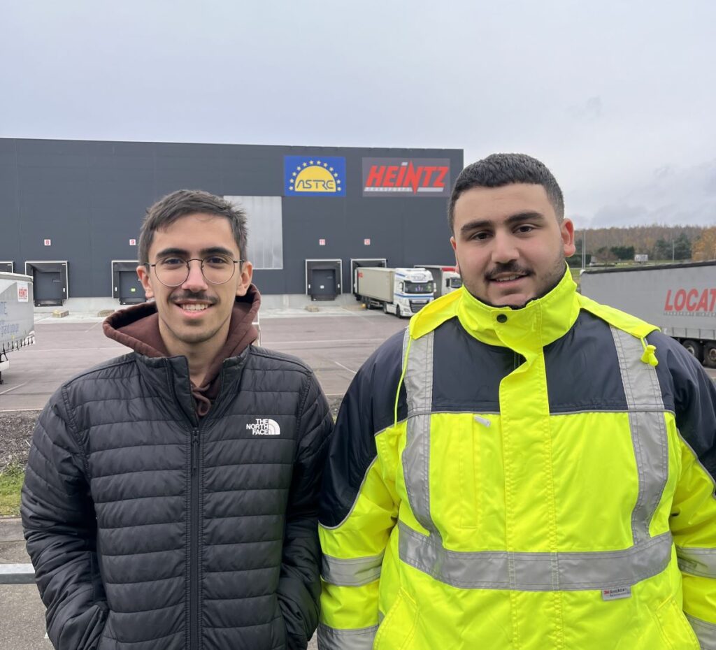 Nos deux alternants Matthieu et Yunus avec le bâtiment logistique en arrière fond